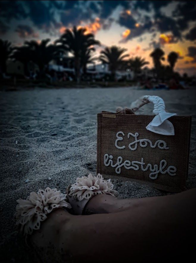 There are legs on a beautiful beach with fine sand and a beautiful summer purse with the inscription EJova lifestyle. There is a sunset with palms in the background.